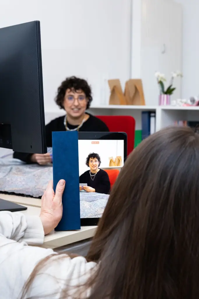 Junge Frau filmt andere junge Frau mit Locken mit einem IPad. Frau mit Locken hält Karten in einer Hand und spricht.