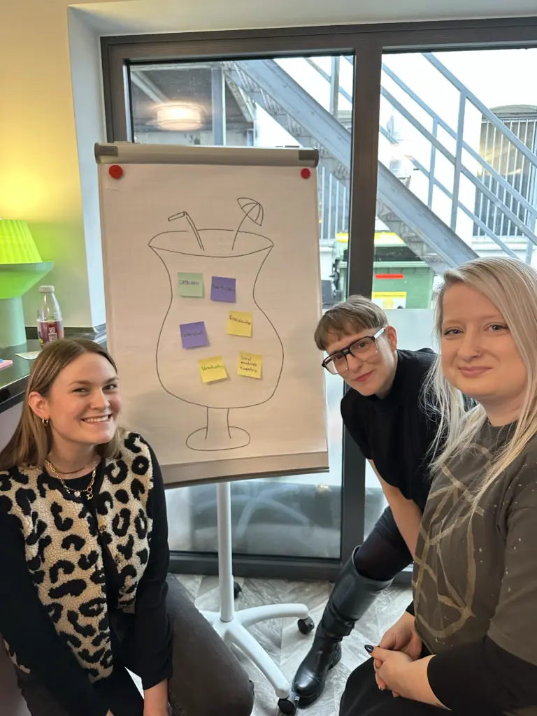Drei Frauen vor einer Flipchart, sie lächeln.