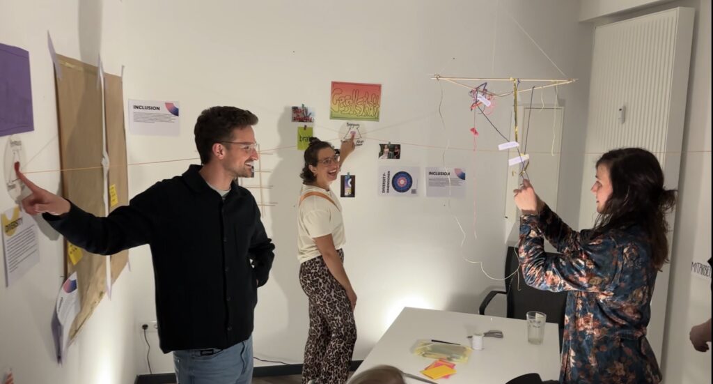Drei Menschen stehen in einem Raum und gestalten eine Ausstellung. Dabei knotet eine Person an einem Mobilee, das von der Decke hängt. Die beiden anderen halten Bänder die vom Mobile ausgehen an der Wand fest. 
