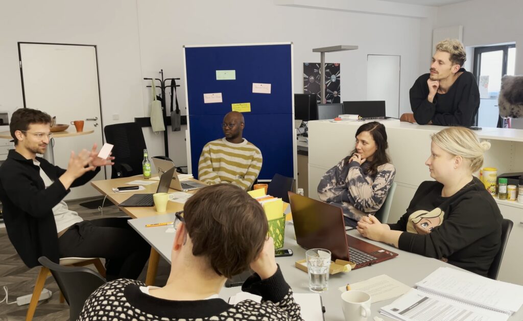 Fünf Kreative sitzen um einen Tisch und diskutieren. Im Hintergrund ist eine Metaplanwand mit einigen Post Its zu sehen. Eine weitere Person hört gespannt zu.